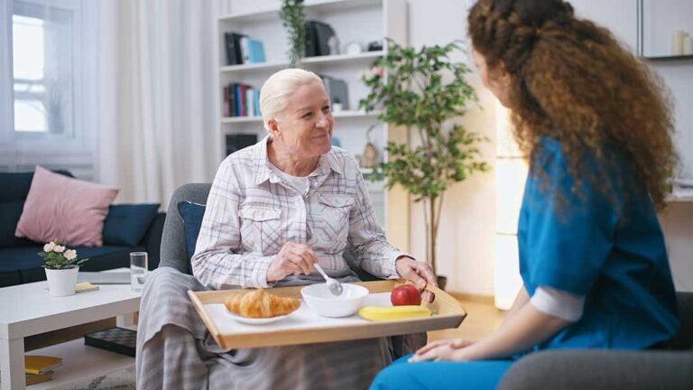 An older adult sits with a younger family member at a kitchen table, talking and looking at photo albums together.
