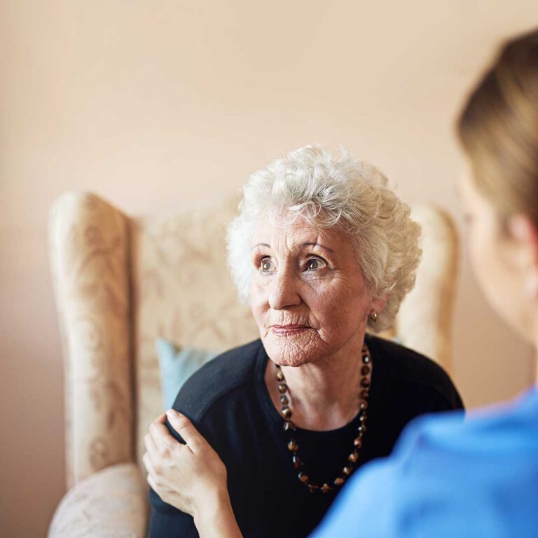 Older adult residents walk together indoors while another resident performs seated stretching nearby.